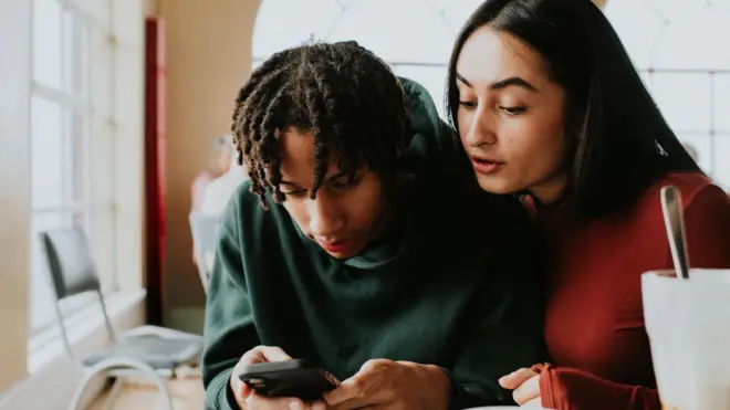 若者2人が、1人が持っているスマートフォンをのぞき込んでいる。スマートフォンを持っている人物は緑色のフーディーを、もう一人の人物は赤い上着を着ている。背景はカフェのような室内。