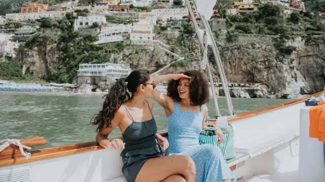 Dos mujeres visiblemente relajadas y felices, disfrutan de un paseo en barco por el mar Mediterráneo. Al fondo se divisa la famosa vista de Positano, Italia.