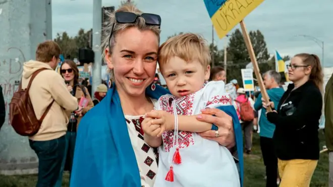 A woman with blonde hair in a Ukrainian flag holds a a boy in her arms
