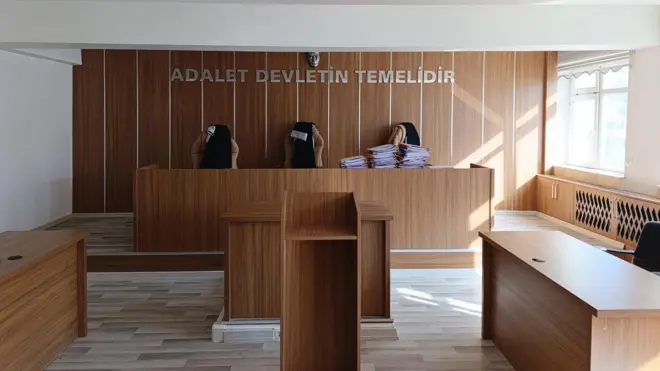 Boş bir mahkeme salonunda, mahkeme heyetinin yer aldığı kürsüde dosyaların yığıldığı görülüyor. 
