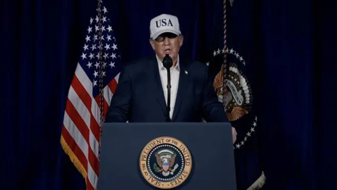 Президент Трамп в костюме и в белой бейсболке с надписью "USA" произносит речь за трибуной