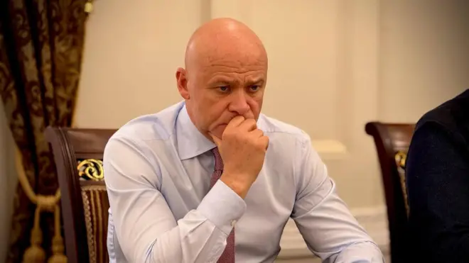 Задумливий Труханов сидить, спершись ліктем на стіл, а другою рукою підпирає підборіддя. Його обличчя серйозне й зосереджене, погляд спрямований вниз. На задньому плані видно частину стільця з різьбленою спинкою та важкі штори.