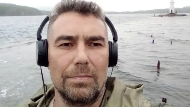 Андрей Акузин в больших черных наушниках фотографирует сам себя на фоне реки и холмов