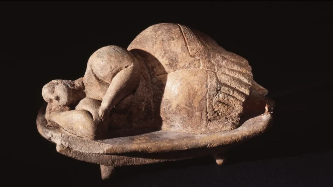 Neolithic clay figurine of a woman sleeping peacefully on her side, one hand under her head, on a simple raised platform. 