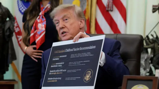 Trump, sentado diante de mesa e com olhar sério, segura cartaz com novas recomendações sobre vacinação infantil.