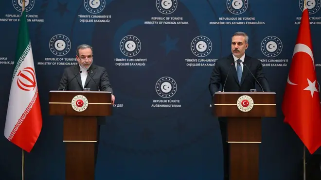 İki erkek kürsülerinin arkasında duruyor. Yanlarında Türkiye ve İran bayrakları var. Arkadaki mavi panoda "Türkiye Cumhuriyeti Dışişleri Bakanlığı" yazıyor. 