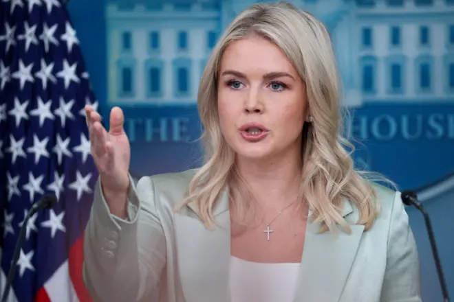 Karoline aponta para a frente enquanto fala em frente a bandeira dos EUA e símbolo da Casa Branca