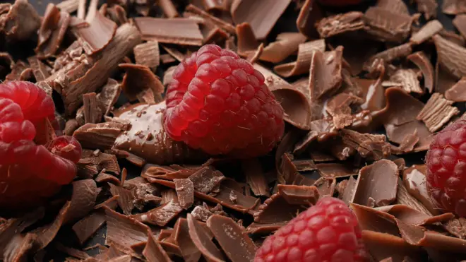 صورة مقربة لرقائق شوكولاتة بنية اللون قرب حبوب من الكاكاو وقليل من سائل الشوكولاتة مع التوت الطازج