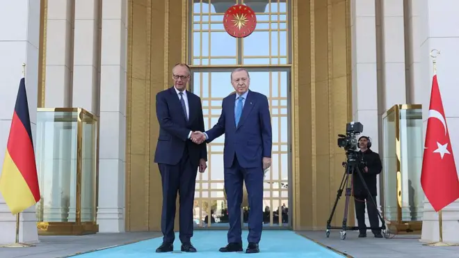 Merz ve Erdoğan Ankara'da Cumhurbaşkanlığında iki ülke bayrakları arasında ayakta tokalaşıyor, arka planda çekim yapan bir kamera var