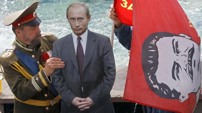 Актер в костюме Николая Второго держит картонную фигуру Путина, а рядом - флаг с портретом Сталина