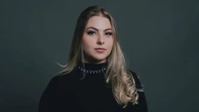 Raphaela Coiado é loira e usa blusa preta e colar prata. Ela olha para a câmera sem sorrir.