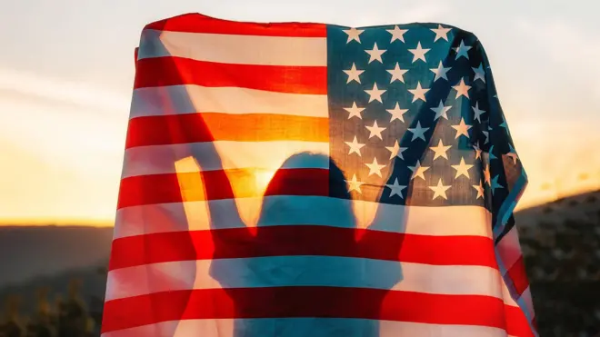 Silueta de una persona con los brazos levantados detrás de una bandera de Estados Unidos iluminada por la luz del atardecer.