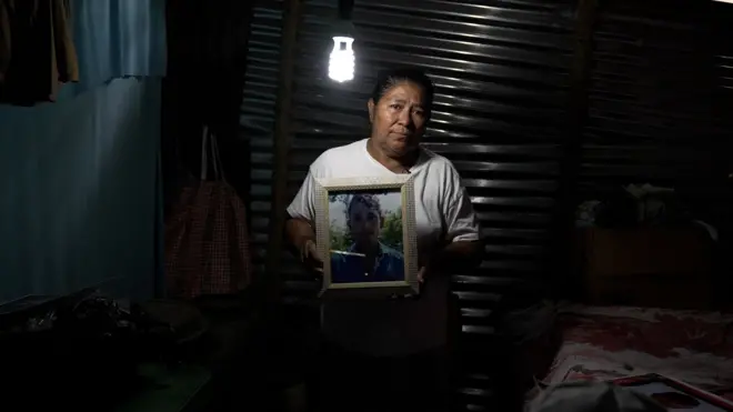 Mulher segura retrato de filho adulto; ela está num quarto de uma casa humilde, à meia luz.