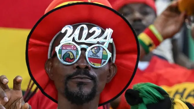 Un supporter de football ghanéen portant des lunettes sur lesquelles est inscrit 2024. Il est coiffé d'un chapeau à bord rouge.