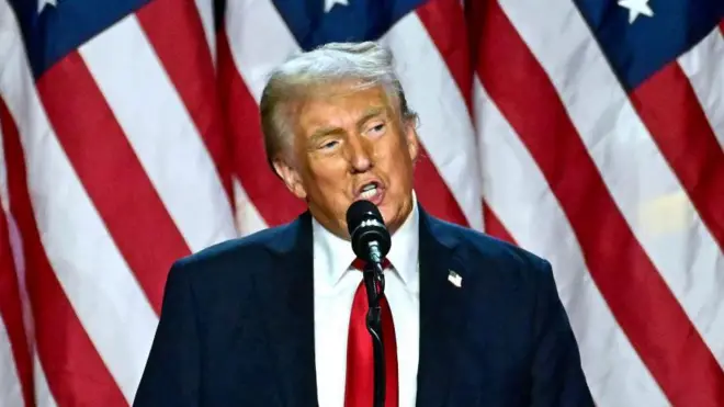 Дональд Трамп говорить у мікрофон, стоячи перед американськими прапорами