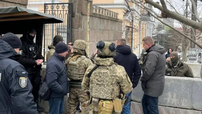 Люди в форме военных и полиции и несколько гражданских стоят у высокого каменного заграждения с металлическими воротами.