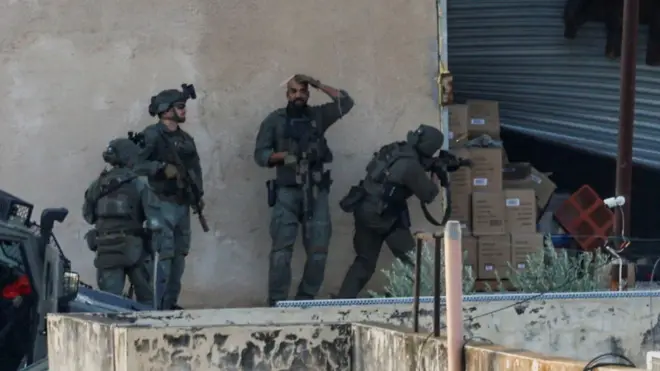 イスラエル兵らが壁の前に立っている。1人はガレージのシャッターの開いた部分から中へと銃を向けている