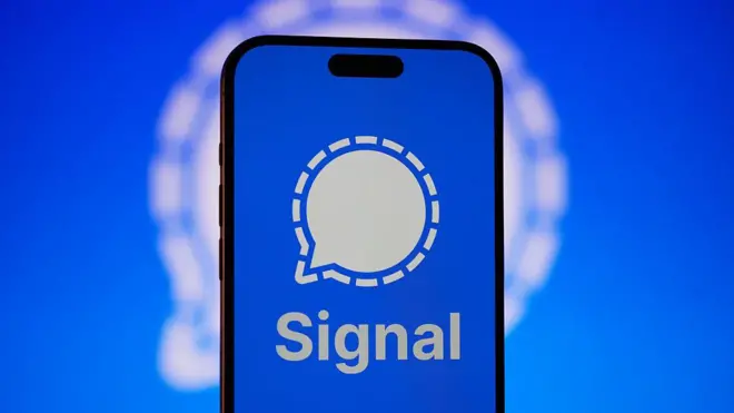一部手機螢幕上顯示Signal應用程式的標誌,背景為藍色,後方則有放大且模糊的 Signal標誌。