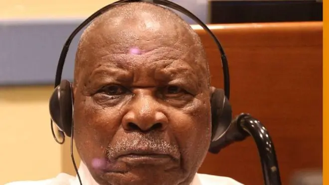 Félicien Kabuga areba muri 'camera' yambaye inyumvisho (écouteurs) mu matwo. Yambaye ishati y'umweru, afite umusatsi mucye w'imvi n'ubwanwa bucye bwo hejuru bw'imvi. 