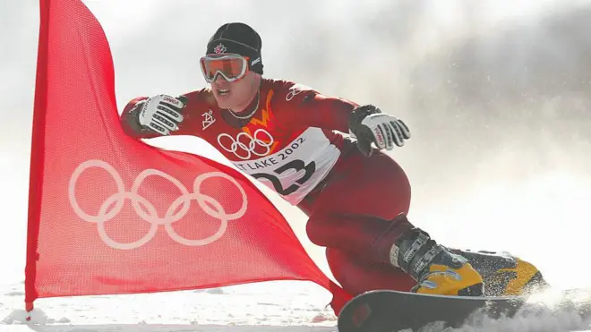 Ryan Wedding en una snowboard con el uniforme rojo de los Juegos Olímpicos de Salt Lake City, gafas de nieve, botas amarillas y un gorro negro. A su lado ondea una bandera roja con el logotipo olímpico en blanco.