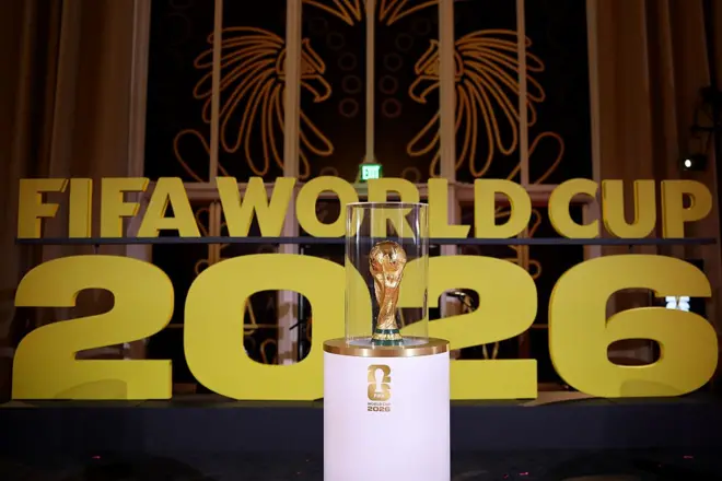 Le trophée de la FIFA entouré de verre transparent, posé sur une table ronde, avec au fond un écrit : "Fifa World Cup 2026" (la Coupe du Monde de la FIFA 2026).