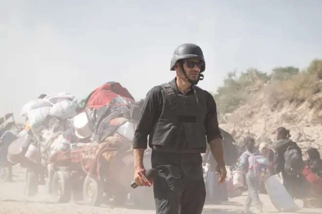 المذيع الإسرائيلي أوهاد حيمو" في تقرير من جباليا شمالي قطاع غزة، يرتدي خوذة ويمسك مايكروفوناً وفي الخلفية صورة النازحين الفلسطينيين