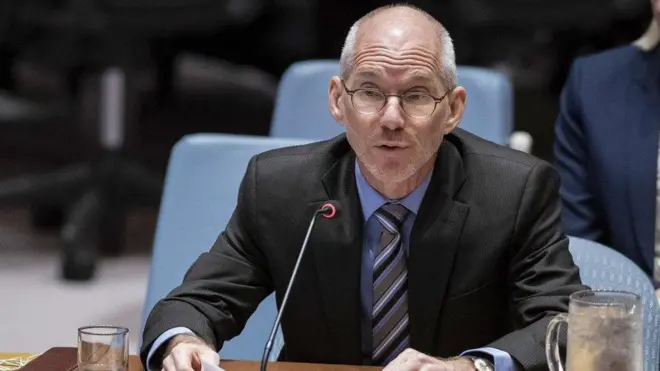 James Swan yambaye ikositimu y'umukara, ishati y'ubururu, na karuvati irimo amabara y'ikigina, ubururu, umweru n'umukara, arimo kuvugira mu ndangururamajwi, iruhande rwe hari ibirahure bibiri ku meza, na telefone igendanwa. 