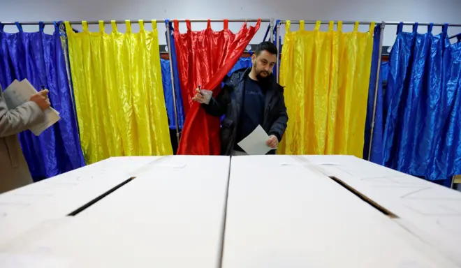 Мужчина выходит из кабинки для голосования с занавесками цветов румынского флага - темно-синими, желтыми и красными - и несет бюллетень к избирательной урне.