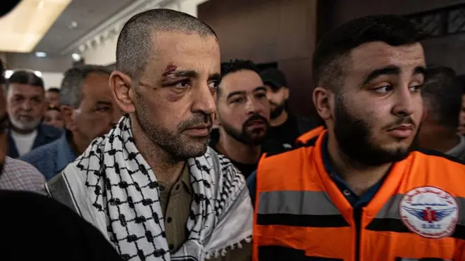 Un palestino que acaba de ser liberado y que tiene una herida alrededor del ojo. Porta una kufiya y va acompañado de un sanitario con un chaleco naranja.