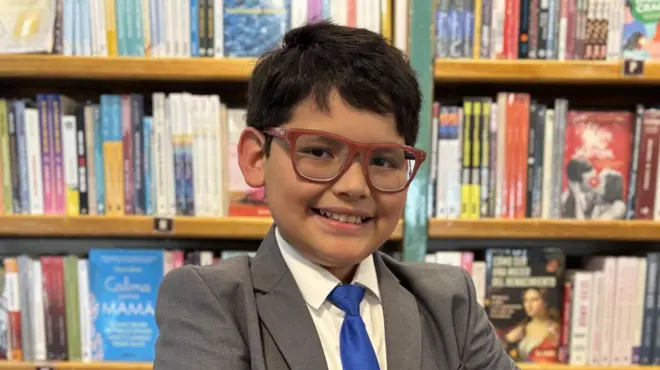 David Camacho, un niño con una sonrisa amplia, anteojos, vestido de saco gris y corbata azul, posa frente al estante de una librería.