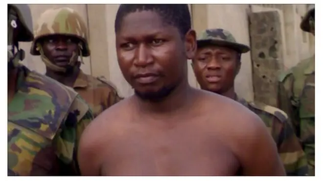 Mohammed Yusuf, bare-chested and with a bandage on his arm, surrounded by soldiers