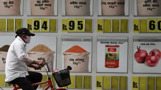 A msn wearign a facemaks cycles past a supermarket with food item prices along a street during a government-imposed nationwide lockdown as a preventive measure against the COVID-19 coronavirus in Colombo on March 30, 2020