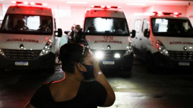 À noite e em frente a três ambulâncias estacionadas, mulher a aparece com a mão no rosto, gesto que remete a preocupação