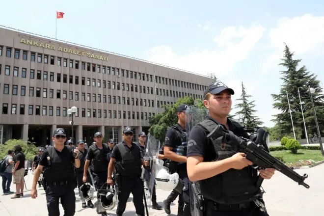 Ankara Adliye binası önünde polisler var. Öndeki polis elindeki silahı kaldırmış. Arkadaki polisler ellerinde kask ve kalkanla yürüyor.