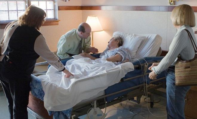 Fotografia colorida mostra uma mulher branca ridosa em um leito de hospital cercada por outras duas mulheres e um homem, todos brancos 