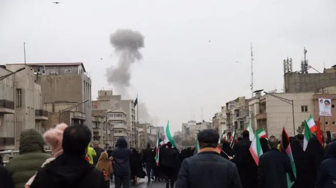 イラン国旗やハメネイ親子の肖像画などを掲げた人たちがカメラに背を向けて、市街地の道路を行進する向こうで、黒煙が上っている