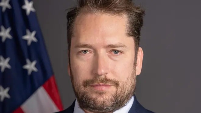 Retrato de Darren Beattie, que aparece sério em frente a bandeira dos EUA