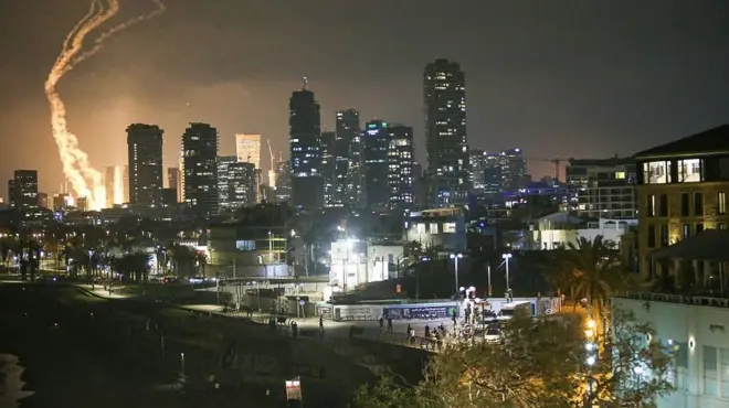 以色列特拉维夫的夜空中，有导弹划过天空的烟雾，远处地面的爆炸造成的火光。