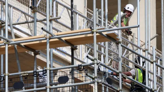 Грађевинска скела и два радника у прслуцима и са кацигама на глави