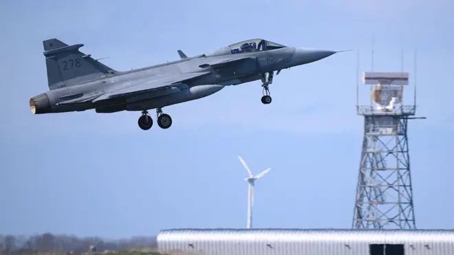 JAS-39 Gripen на взлете: нос самолета направлен по диагонали вверх, за ним видны белые лопасти ветряной электростанции и решетчатая башня аэродрома с антеннами