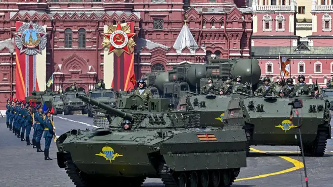 На Червоній площі рухається колона бронетехніки кольору, зокрема гусеничні бойові машини з військовослужбовцями всередині та на броні. Ліворуч видно підрозділ військових у парадній формі, які стоять шеренгою. На тлі – великі історичні будівлі з червоної цегли, прикрашені святковими банерами та емблемами.