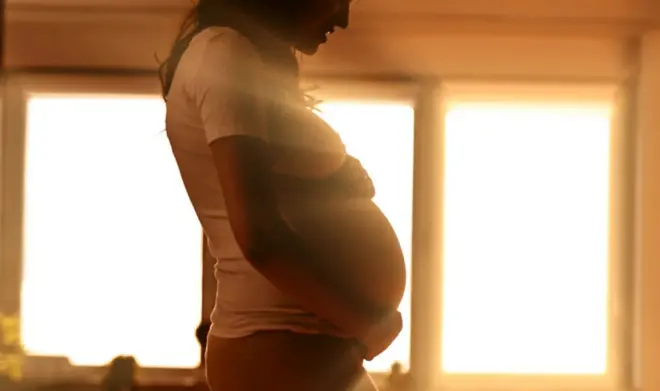 Pregnant woman standing in front of brightly backlit window Umugore utwite ahagaze imbere y'idirishya ahaturuka urumuri inyuma ye.