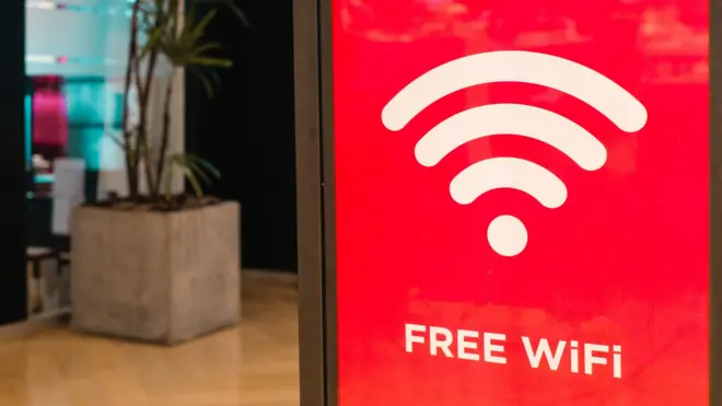 Cartaz vermelho oferecendo wifi grátis em um ambiente que parece ser o lobby de um hotel