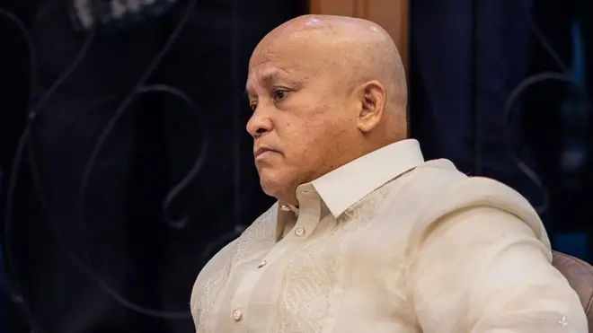 Ronald Dela Rosa, seorang pria berkepala botak mengenakan kemeja berkancing berwarna krem, duduk dan menatap ke kejauhan.