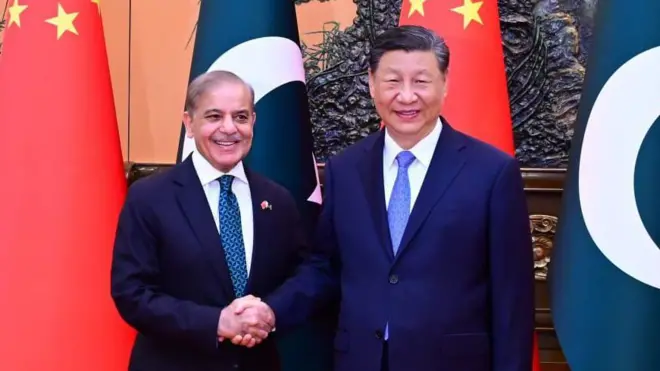 Pakistani President Shehbaz Sharif shakes hands with Chinese President Xi Jinping as the leaders smile while standing in front of the national flags of China and Pakistan.