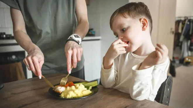 Criança de cabelo castanho com camisa branca, comendo um tomate, com uma mulher vestindo uma camiseta verde cortando um prato de comida na sua frente
