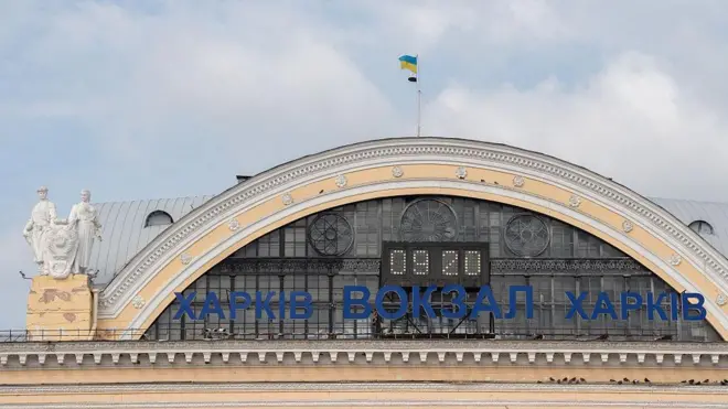 Верхня частина будівлі харківського вокзалу: великий арковий фасад із декоративними елементами та годинником посередині. На даху майорить прапор України. Уздовж арки розміщено напис «ХАРКІВ ВОКЗАЛ ХАРКІВ» синіми літерами.