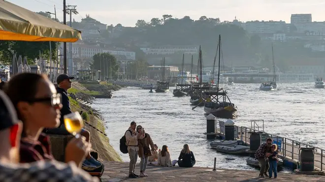 Zachód słońca w Porto na północy Portugalii. Ludzie siedzą nad brzegiem rzeki Douro. Jedna z kobiet, na pierwszym planie po lewej, trzyma kieliszek wina. Inni przechadzają się nabrzeżem i robią zdjęcia.