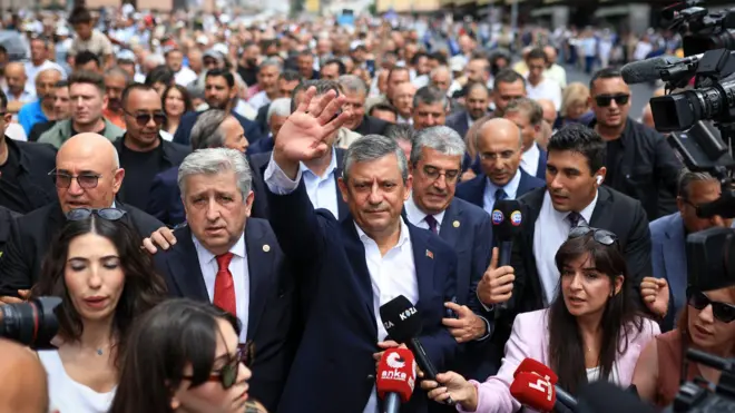 Özgür Özel Hacı Bayram Veli Camii'den çıkıp 1. TBMM binasına yürürken arkasında geniş bir kalabalık olduğu ve sağ elini sallayarak vatandaşları selamladığı görülüyor.