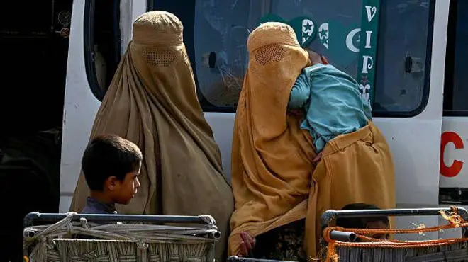 تصویر دو زن مهاجر با دو کودک. یکی از این زنان چادری زرد به سر دارد و کودکش را در آغوش گرفته. دیگری چادری قهوه‌یی به سر دارد و یک پسر بچه در کنارش ایستاده. آن‌ها سمت دوربین نگاه میکنند اما از زیر چادری چهره‌شان معلوم نیست. 